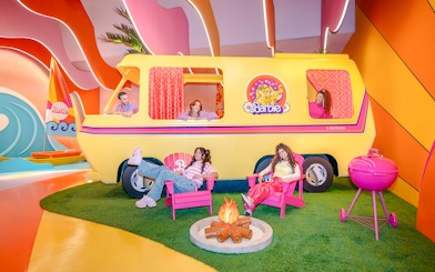 Barbie Malibu van with people relaxing in chairs and a campfire nearby.