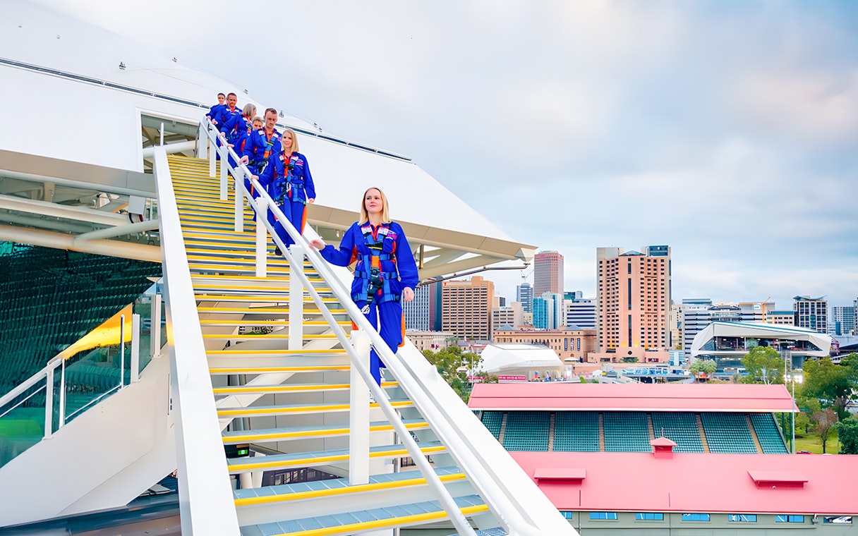 Group on guided tour atop Adelaide Oval Stadium rooftop with city skyline.