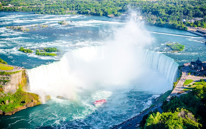 Niagara Falls with tour boat in summer, surrounded by lush greenery.