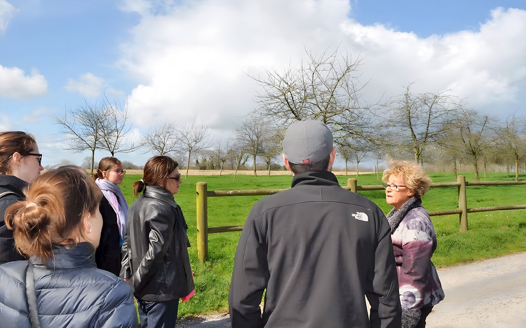 Tour group listening to a guide at Normandy, France, with green fields and trees in the background.