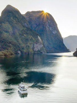 Cruise boat on Doubtful Sound with sun peeking over lush mountains, New Zealand.