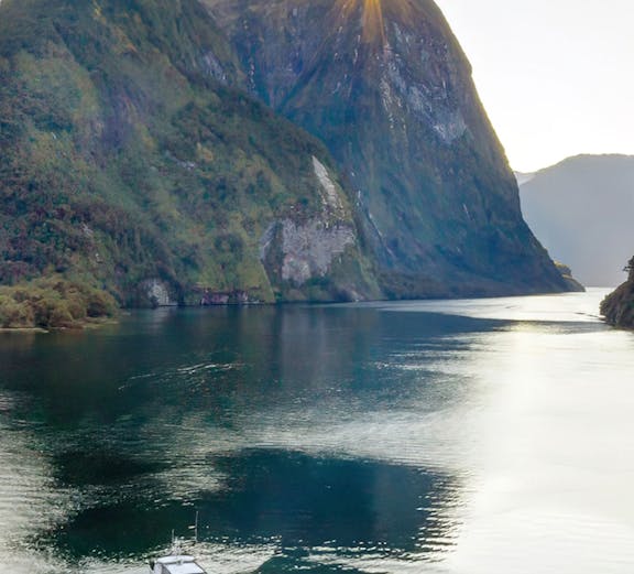 Cruise boat on Doubtful Sound with sun peeking over lush mountains, New Zealand.