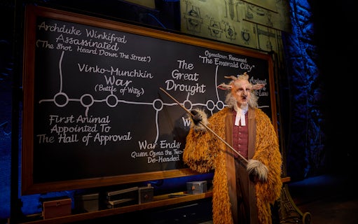 Professor in costume pointing at a chalkboard with historical events from the musical Wicked.