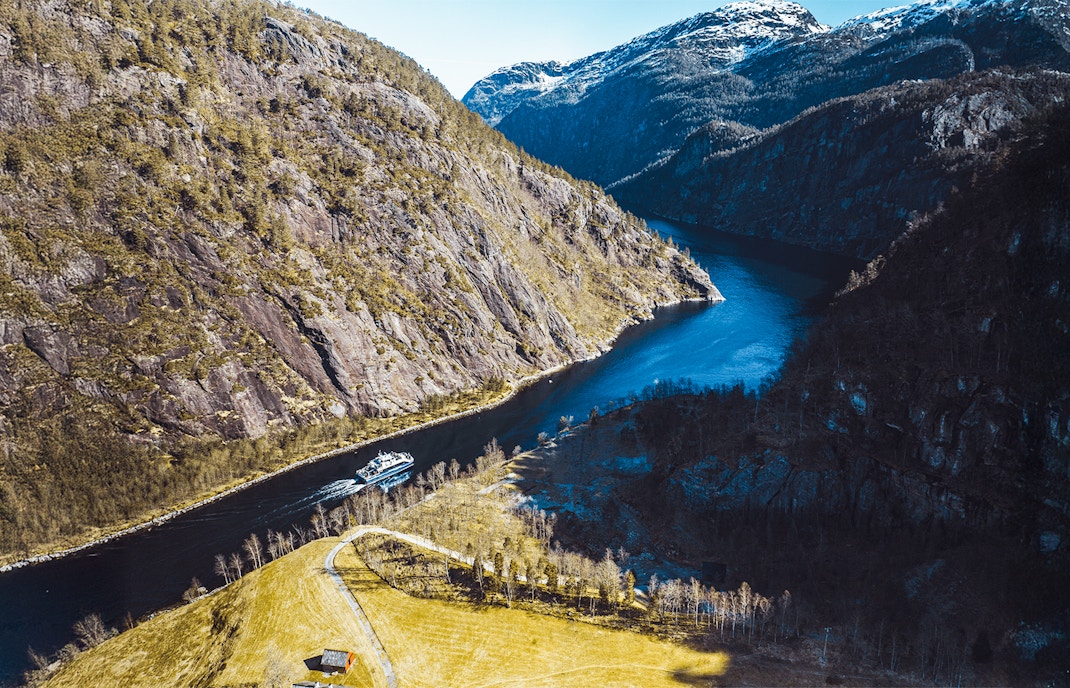 Aerial view of a cruise ship navigating Mostraumen Fjord, surrounded by steep cliffs and blue water.