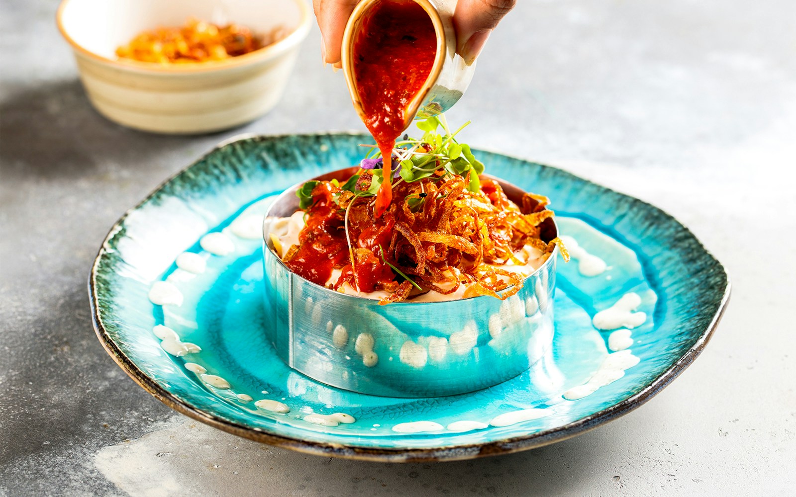 Pouring red sauce over a dish with crispy onions on a blue plate.