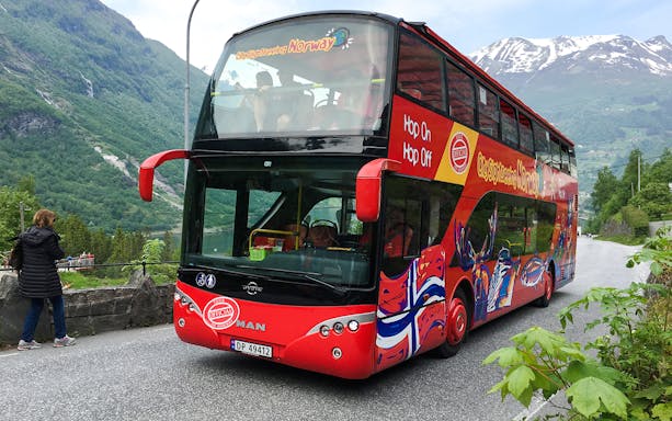 Red double-decker bus on Geiranger Hop-on Hop-off tour with mountains in the background.