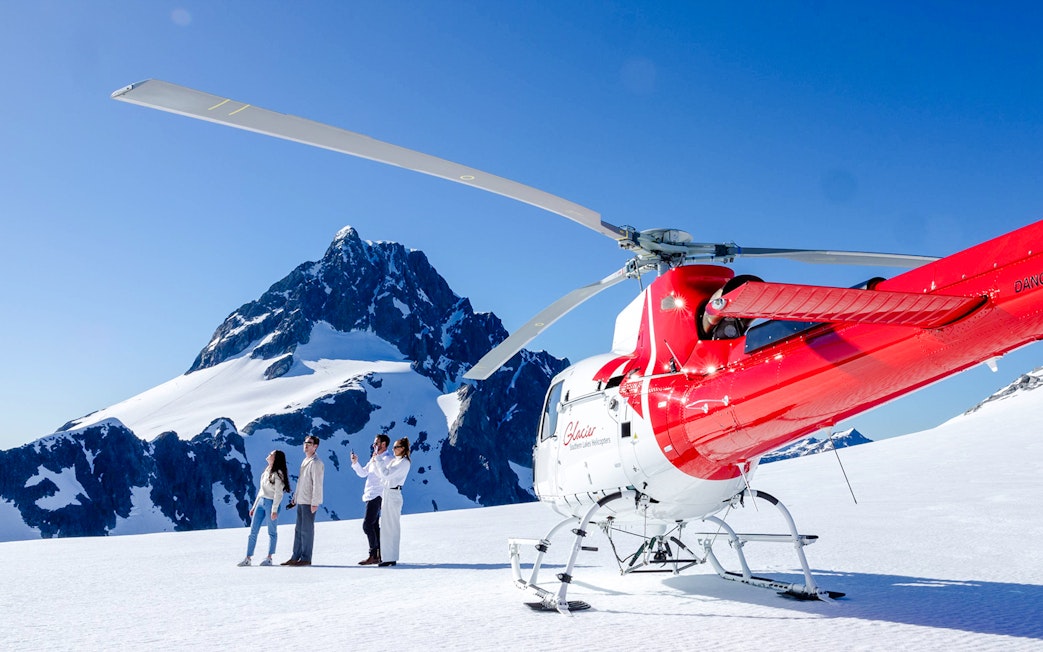Four people beside a red and white helicopter on a snowy peak in Milford Sound.