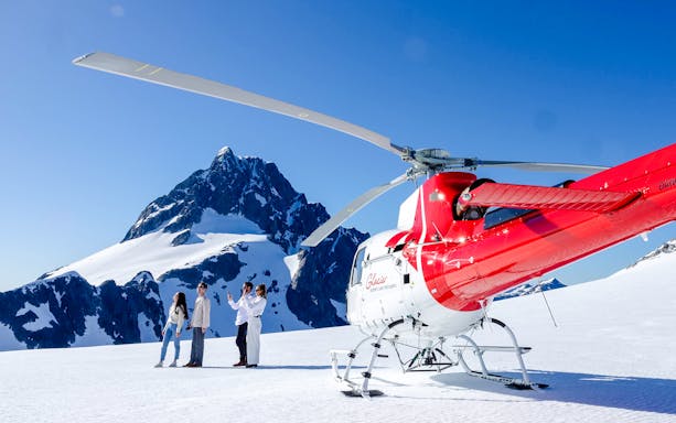 Three people beside a red and white helicopter on a snowy peak in Milford Sound.