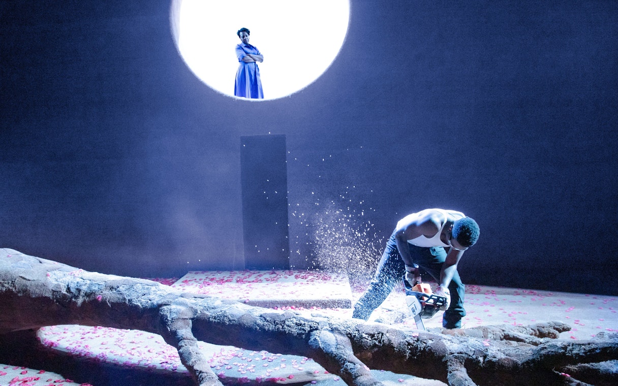 Stage scene from All My Sons West End show with actor using chainsaw on tree trunk.