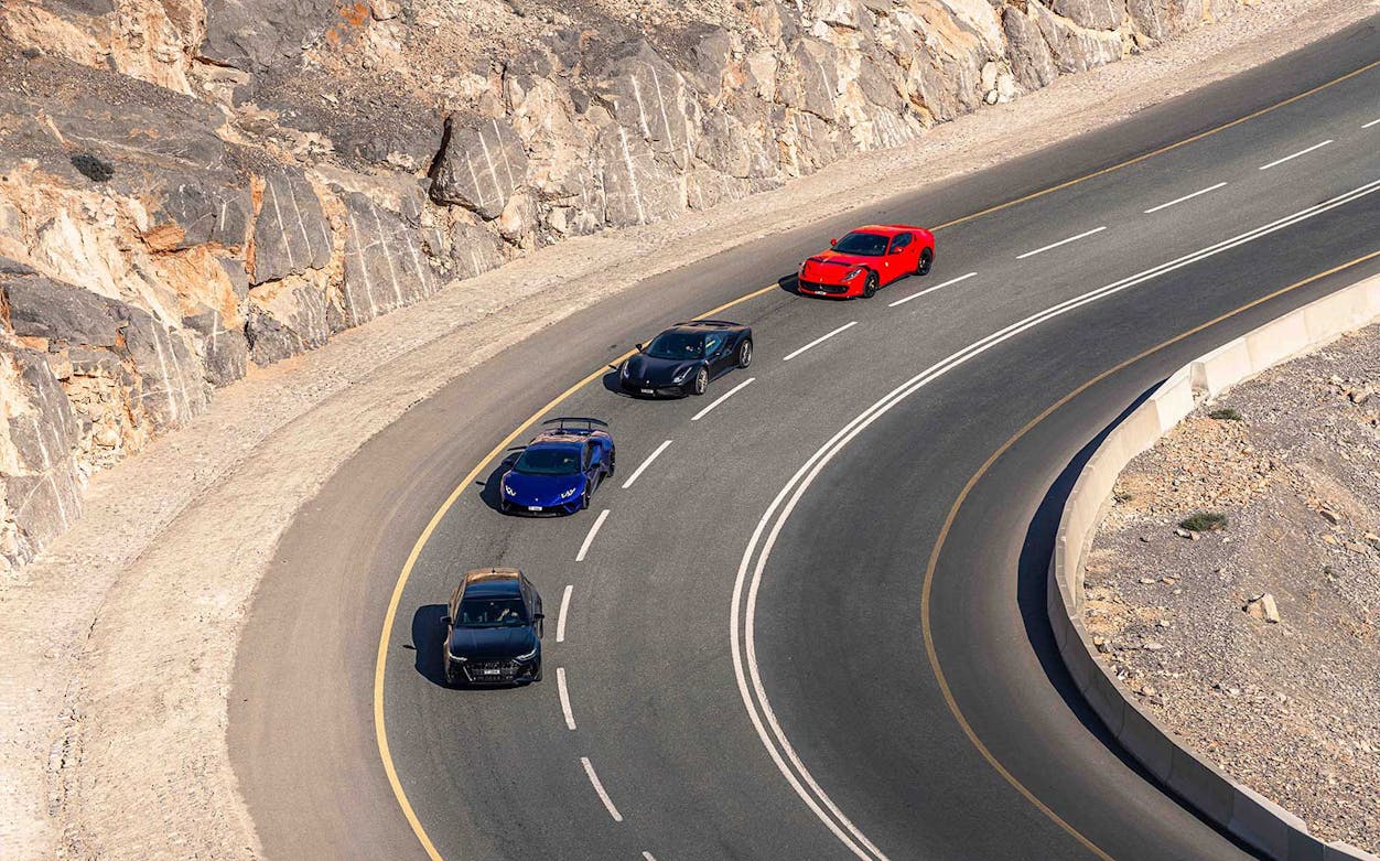 Supercars driving on a winding road at Jabal Bil Ays, surrounded by rocky terrain.