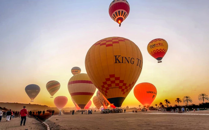 Hot air balloons ascending at sunrise in Luxor, Egypt, with people preparing for a ride.