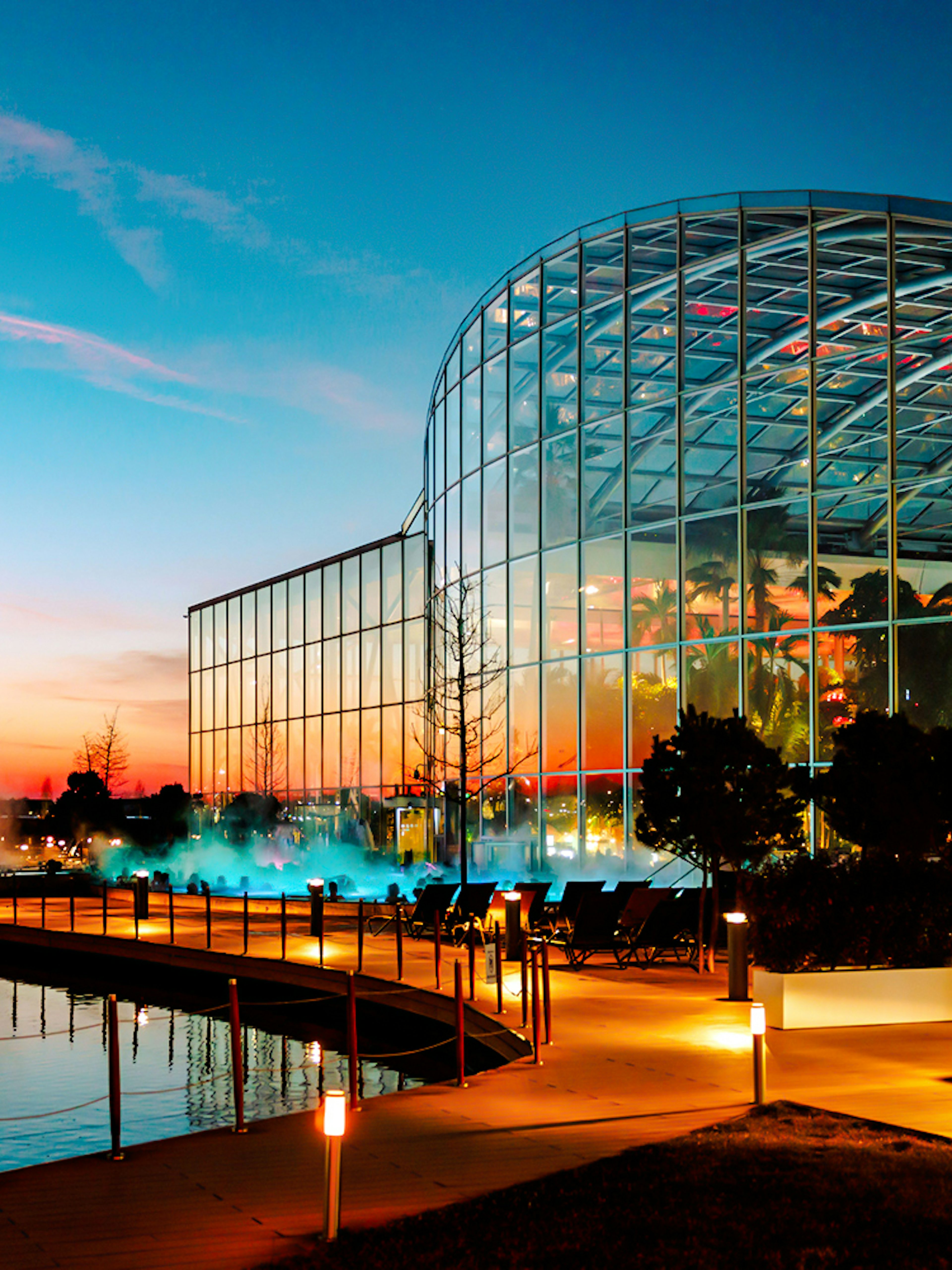 Therme Bucharest illuminated at night with glass facade reflecting sunset colors.