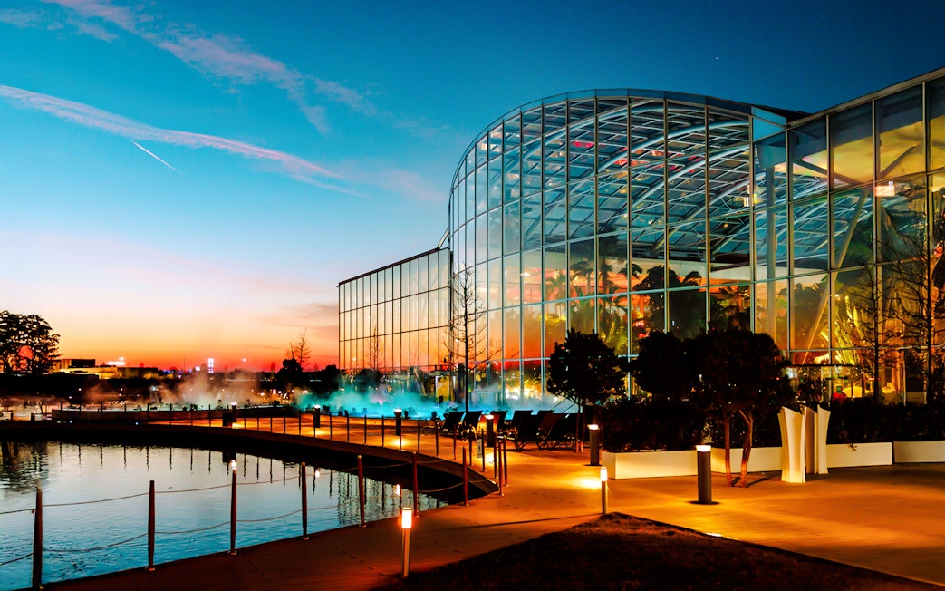 Therme Bucharest illuminated at night with glass facade reflecting sunset colors.