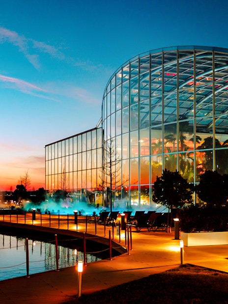 Therme Bucharest illuminated at night with glass facade reflecting sunset colors.