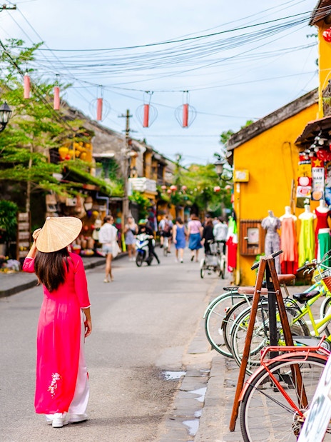 Street scene in Hoi An Ancient Town, Vietnam, with a woman in traditional dress and colorful shops.