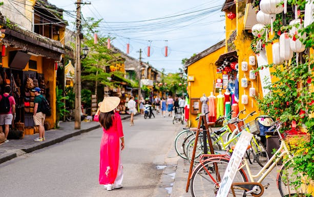 Street scene in Hoi An Ancient Town, Vietnam, with a woman in traditional dress and colorful shops.