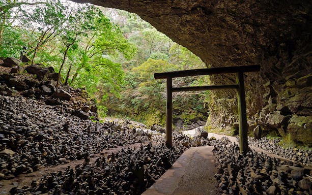 Torii gate at the entrance of a cave in Takachiho, surrounded by stacked stones and lush greenery.