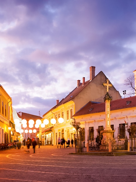 Old town square of Szentendre at dusk with illuminated buildings and a central cross monument.
