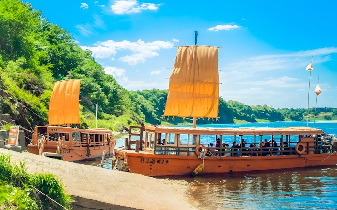Traditional boats with orange sails docked by a river in a lush green landscape, DMZ tour.