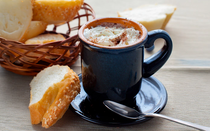 Hot chocolate with whipped cream in a mug beside a basket of sliced baguette.