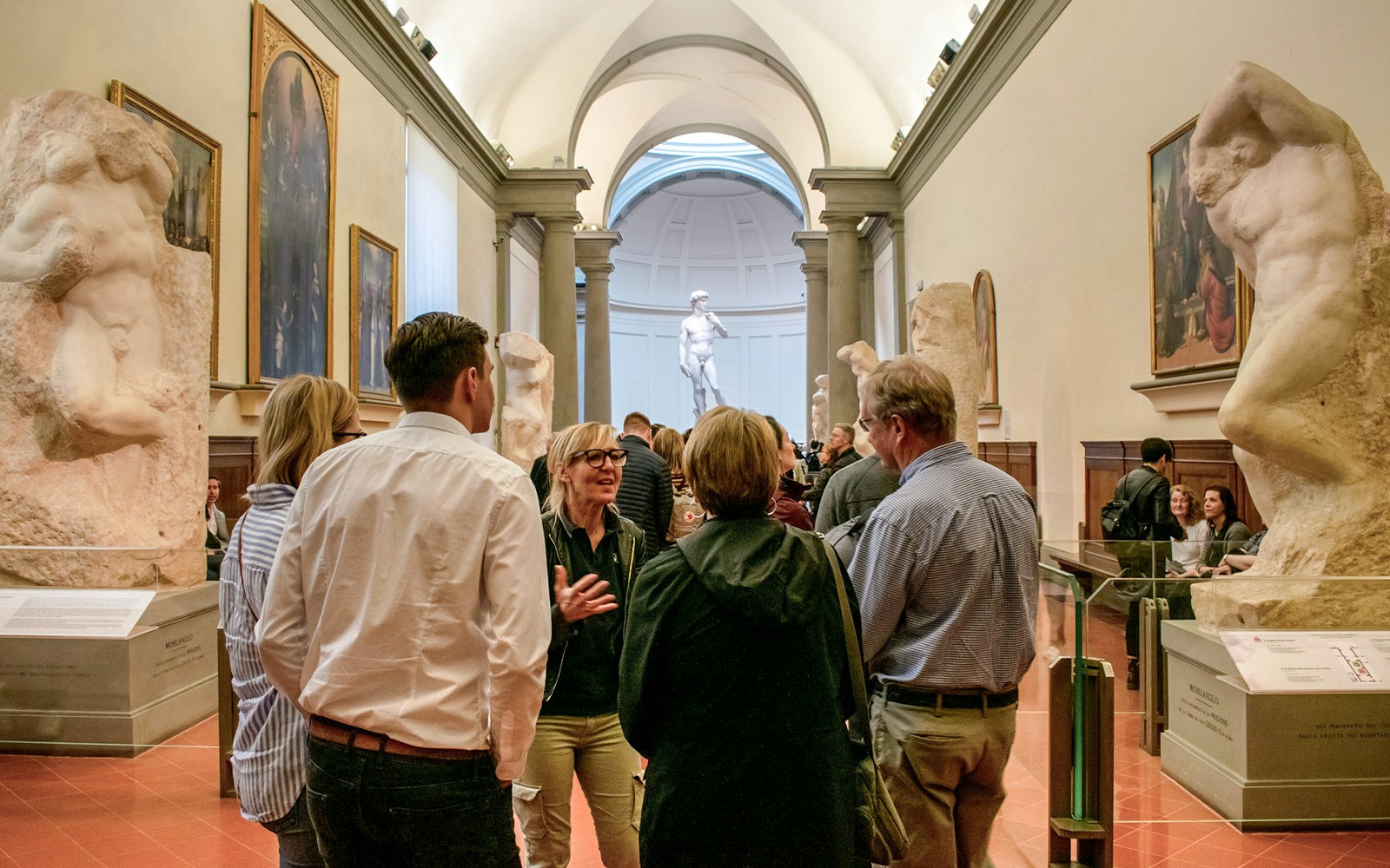 Guide leading tour group in Accademia Gallery, Florence, with Michelangelo's David in background.