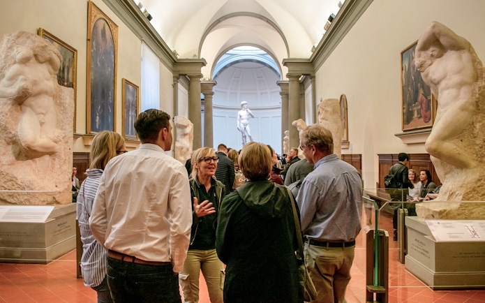 Guide leading tour group in Accademia Gallery, Florence, with Michelangelo's David in background.