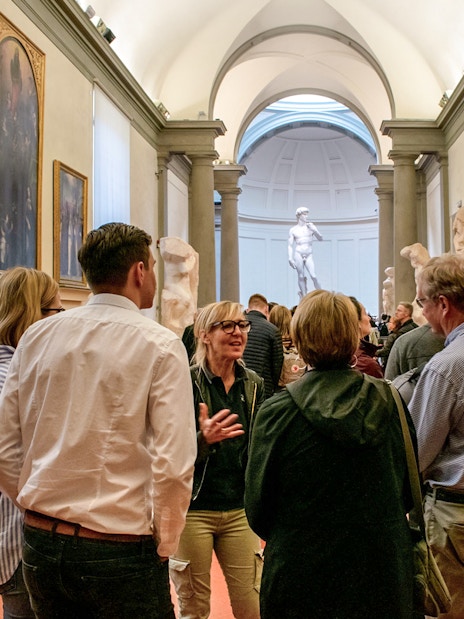 Guide leading tour group in Accademia Gallery, Florence, with Michelangelo's David in background.