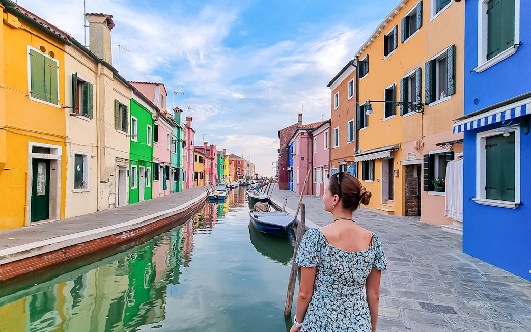 Tourist woman with hat viewing colorful houses along canal in Burano, Italy.