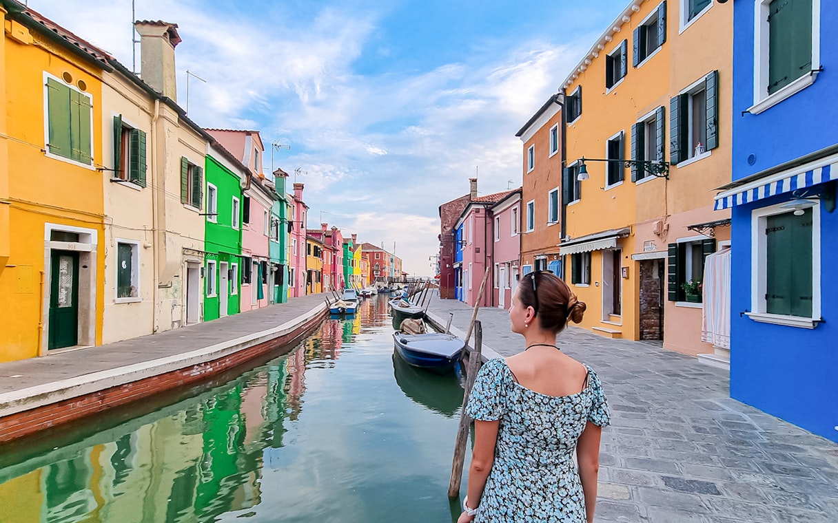 Tourist woman with hat viewing colorful houses along canal in Burano, Italy.