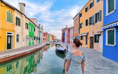 Tourist woman with hat viewing colorful houses along canal in Burano, Italy.
