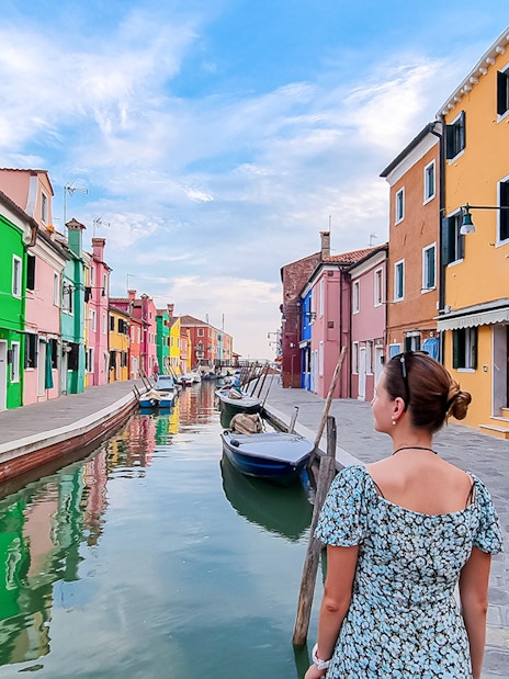 Tourist woman with hat viewing colorful houses along canal in Burano, Italy.
