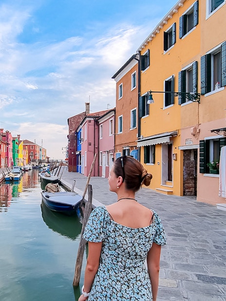 Tourist woman with hat viewing colorful houses along canal in Burano, Italy.
