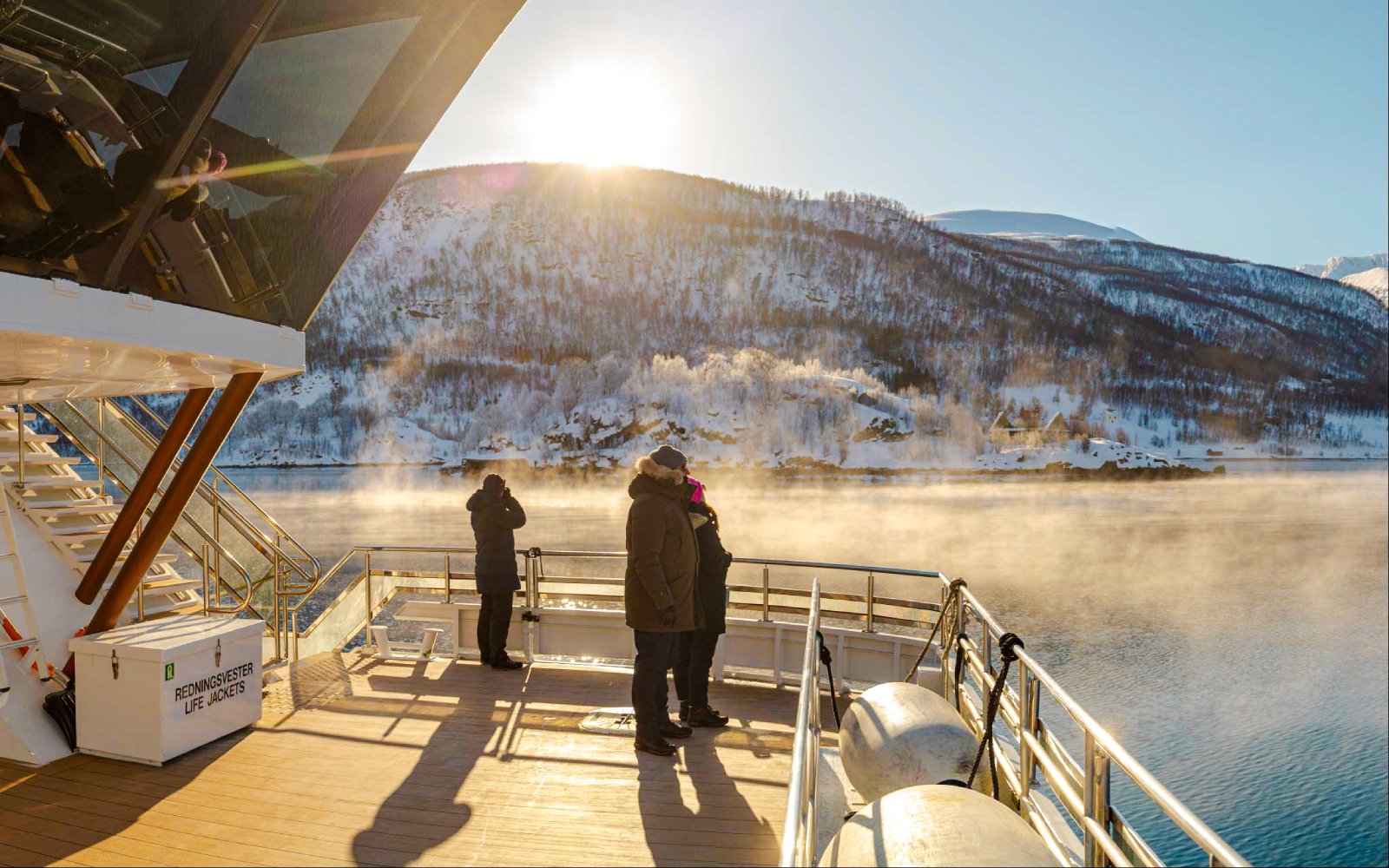 Guests on Arctic Fjord Cruise viewing snowy mountains and misty water in sunlight.