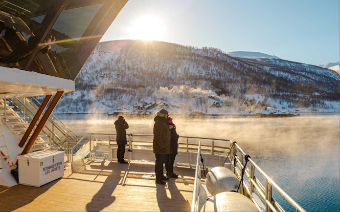 Guests on Arctic Fjord Cruise viewing snowy mountains and misty water in sunlight.