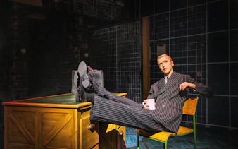 Performer in pinstripe suit with feet on desk during Operation Mincemeat, Broadway show.
