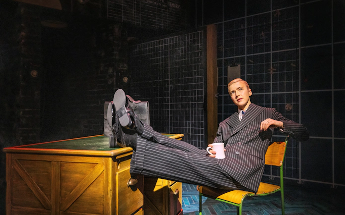 Performer in pinstripe suit with feet on desk during Operation Mincemeat, Broadway show.