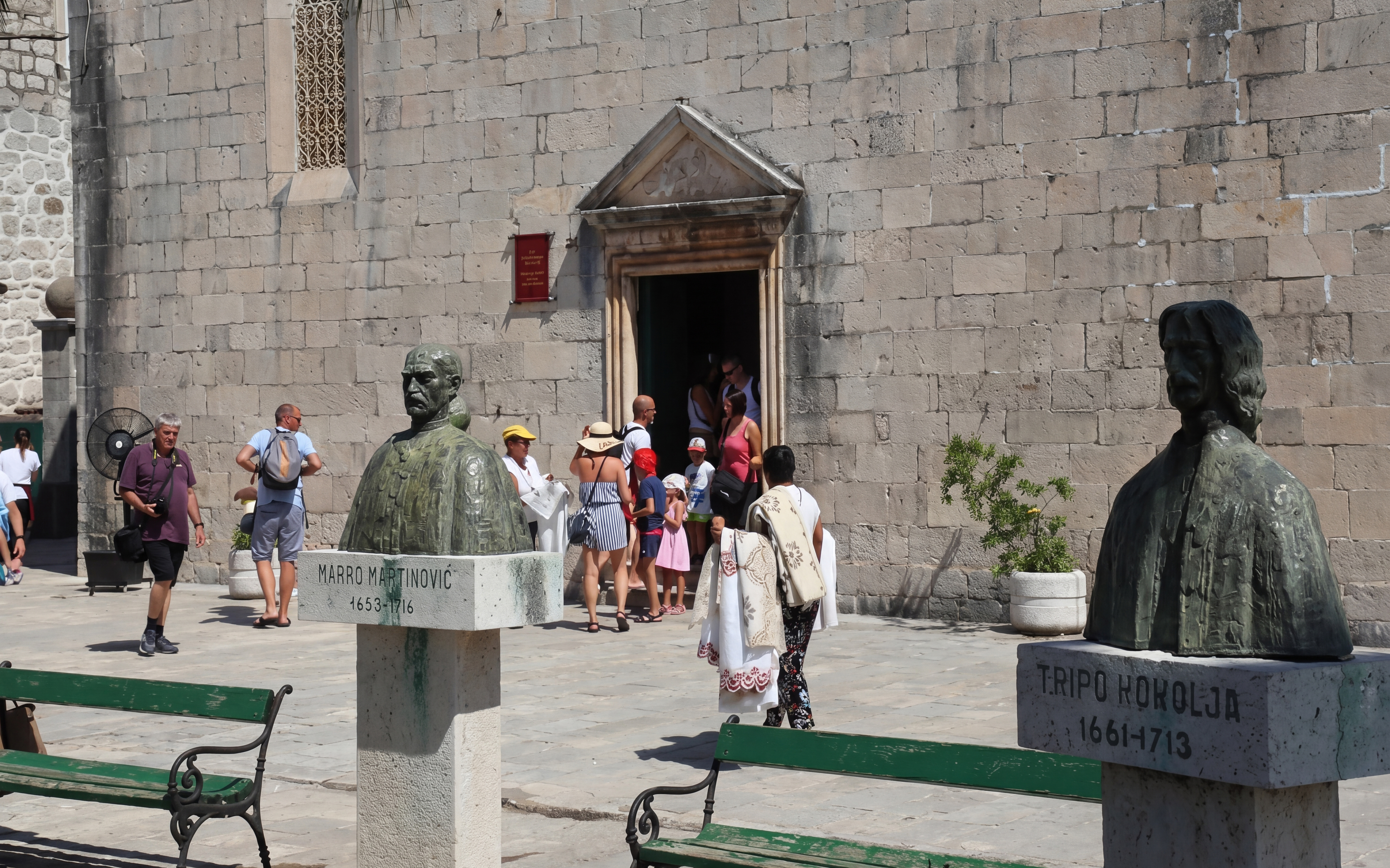 Busts of historical figures in Perast Old Town with tourists entering a stone building.