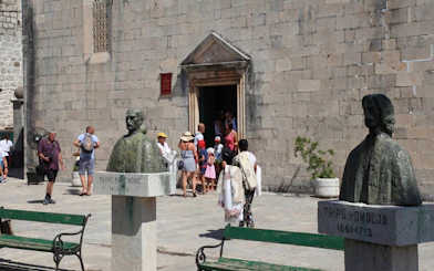 Busts of historical figures in Perast Old Town with tourists entering a stone building.