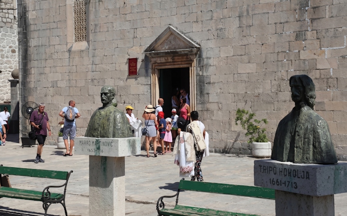 Busts of historical figures in Perast Old Town with tourists entering a stone building.