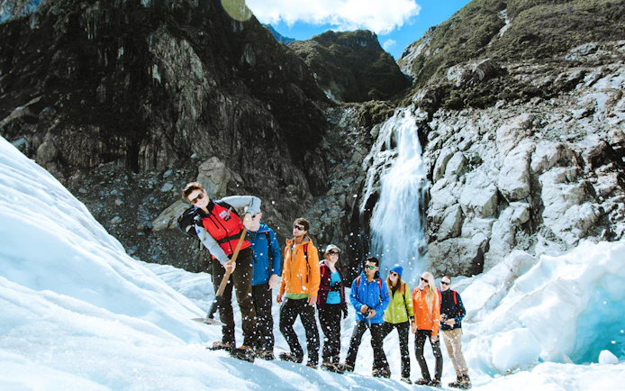 Tour group hiking on Fox Glacier, New Zealand with a waterfall in the background.