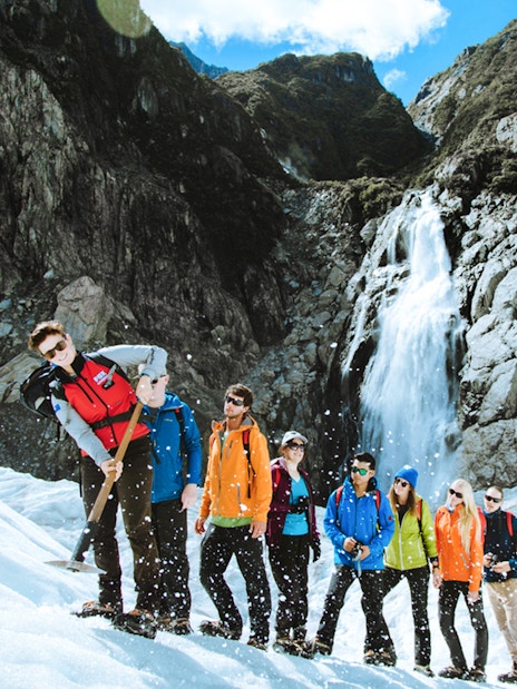 Tour group hiking on Fox Glacier, New Zealand with a waterfall in the background.