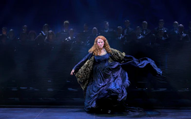 A performer in a dramatic scene from My Fair Lady, with a choir in the background.