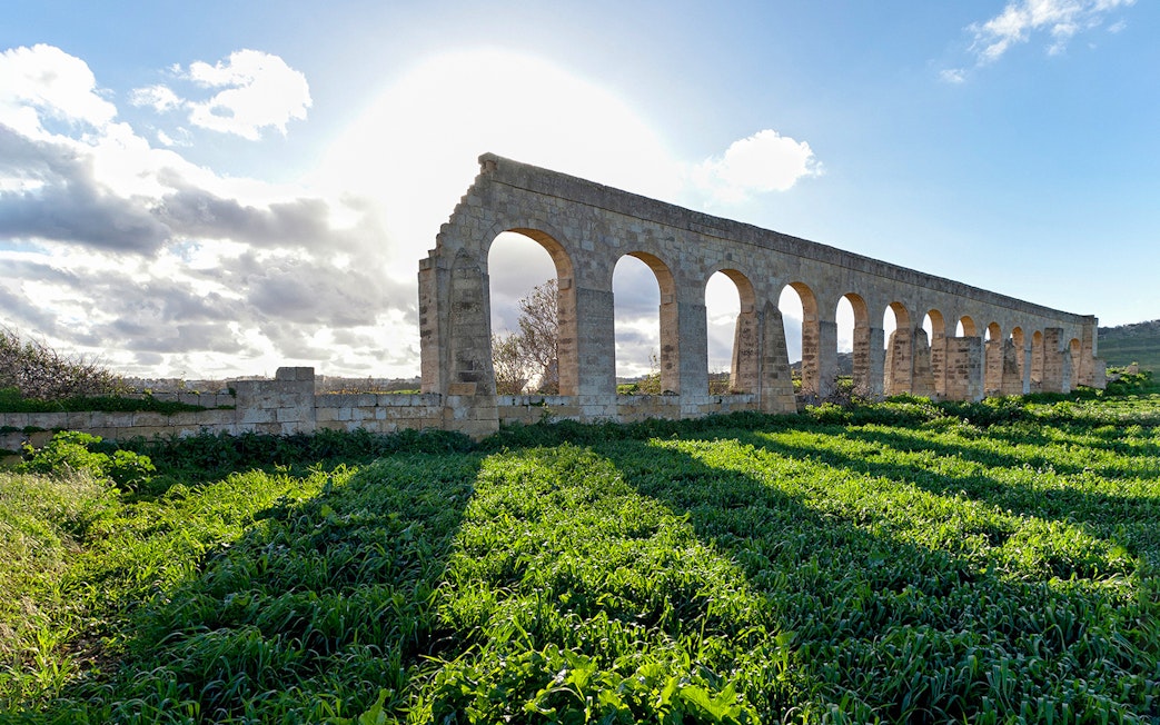 Ancient stone aqueducts on Gozo Island with green fields and a bright sky.