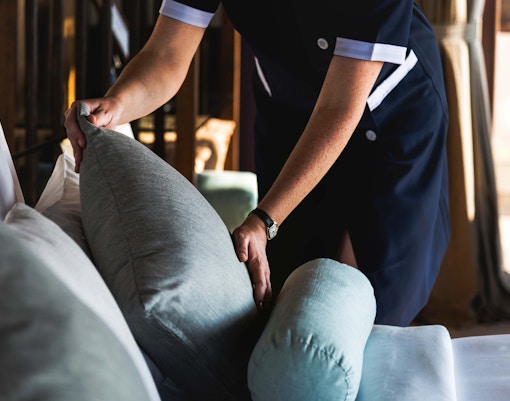 The image shows a person, likely a hotel worker or housekeeper, arranging pillows on a bed or sofa.
