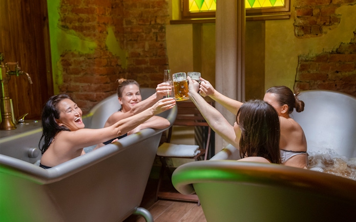 Four women enjoying a beer spa experience, toasting with drinks in metal tubs.
