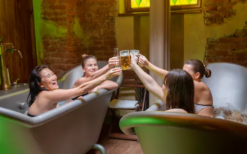 Four women enjoying a beer spa experience, toasting with drinks in metal tubs.