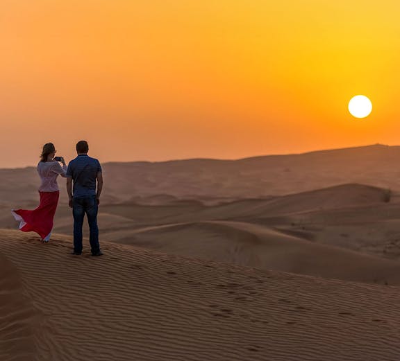 Couple standing on sand dune at sunset during desert safari.