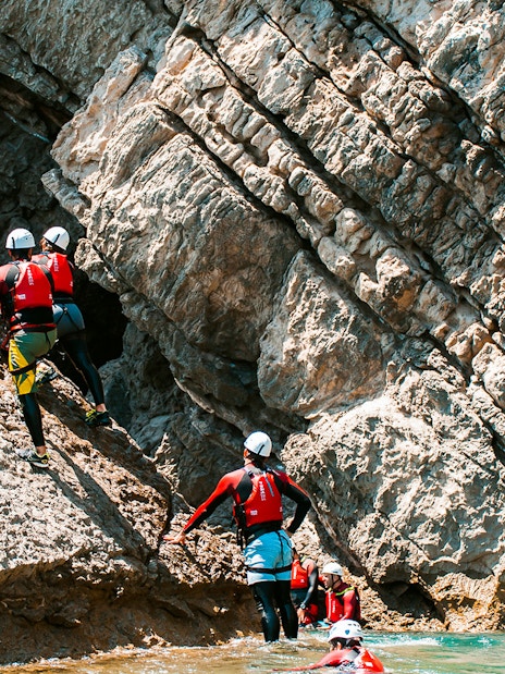 Group coasteering along rocky cliffs in wetsuits and helmets.