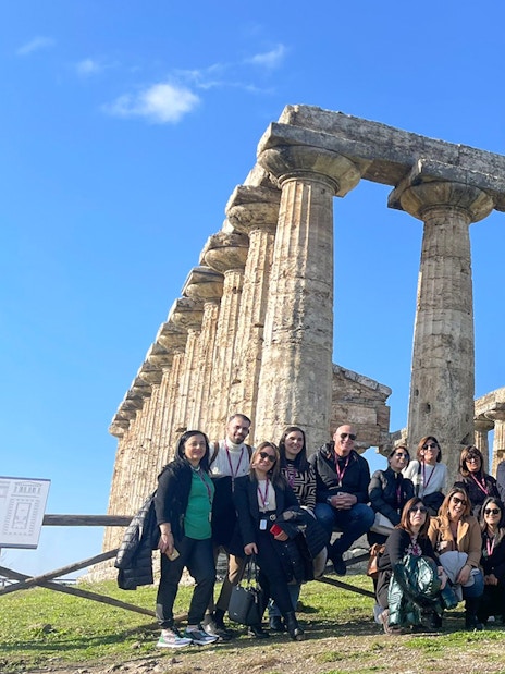 Tour group with guide at Paestum Archaeological Site, Italy, in front of ancient temple ruins.