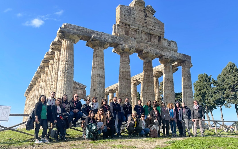 Tour group with guide at Paestum Archaeological Site, Italy, in front of ancient temple ruins.